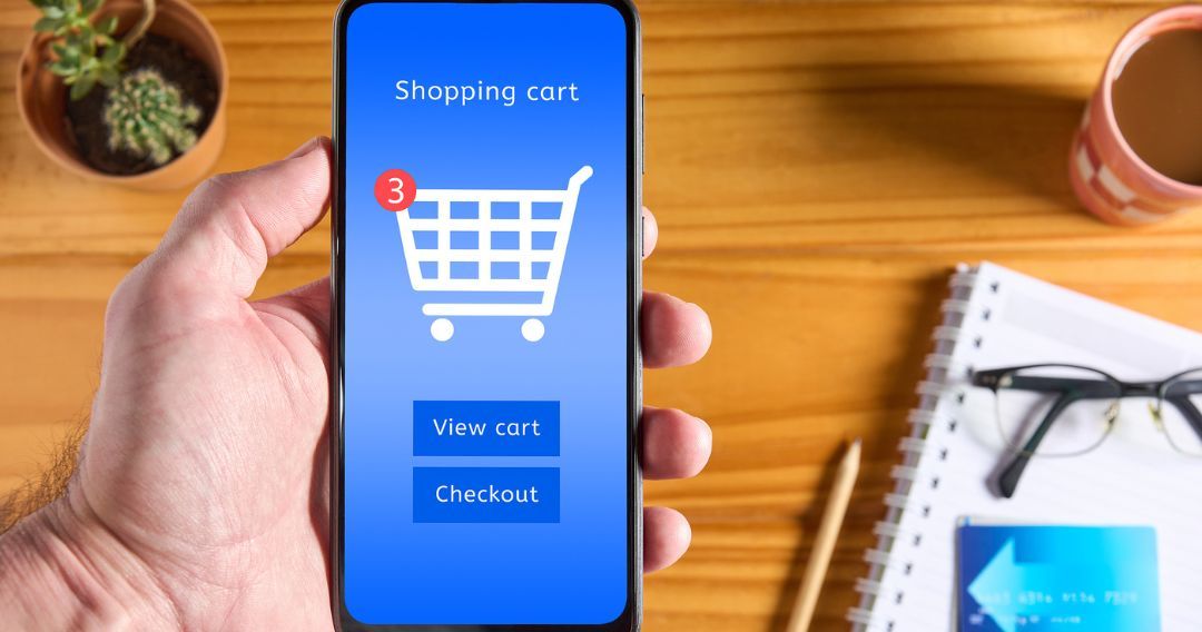 Male hand holding mobile phone with interface of online store on the screen, shopping cart icon and buttons to view cart and checkout. Credit card and accessories on table in the background